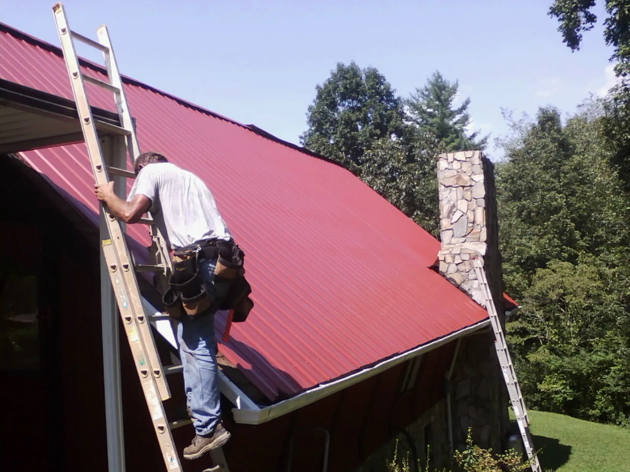 Roofer on a ladder beside a red metal roof and stone chimney — Soffit & Fascia Repair by Roofing Brothers in Machipongo